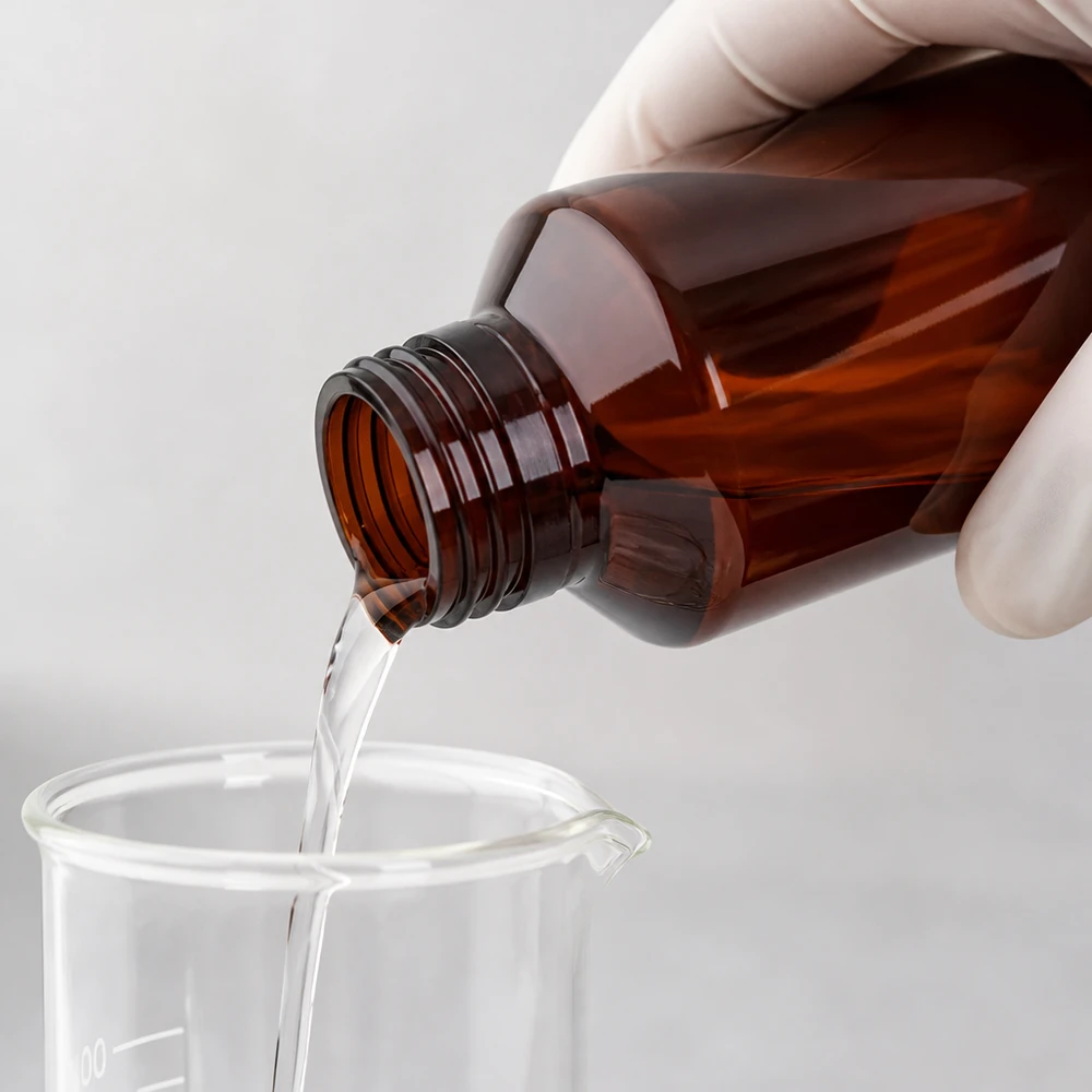 Een met een handschoen beschermde hand houdt een bruine plastic fles van 100 ml boven een maatbeker