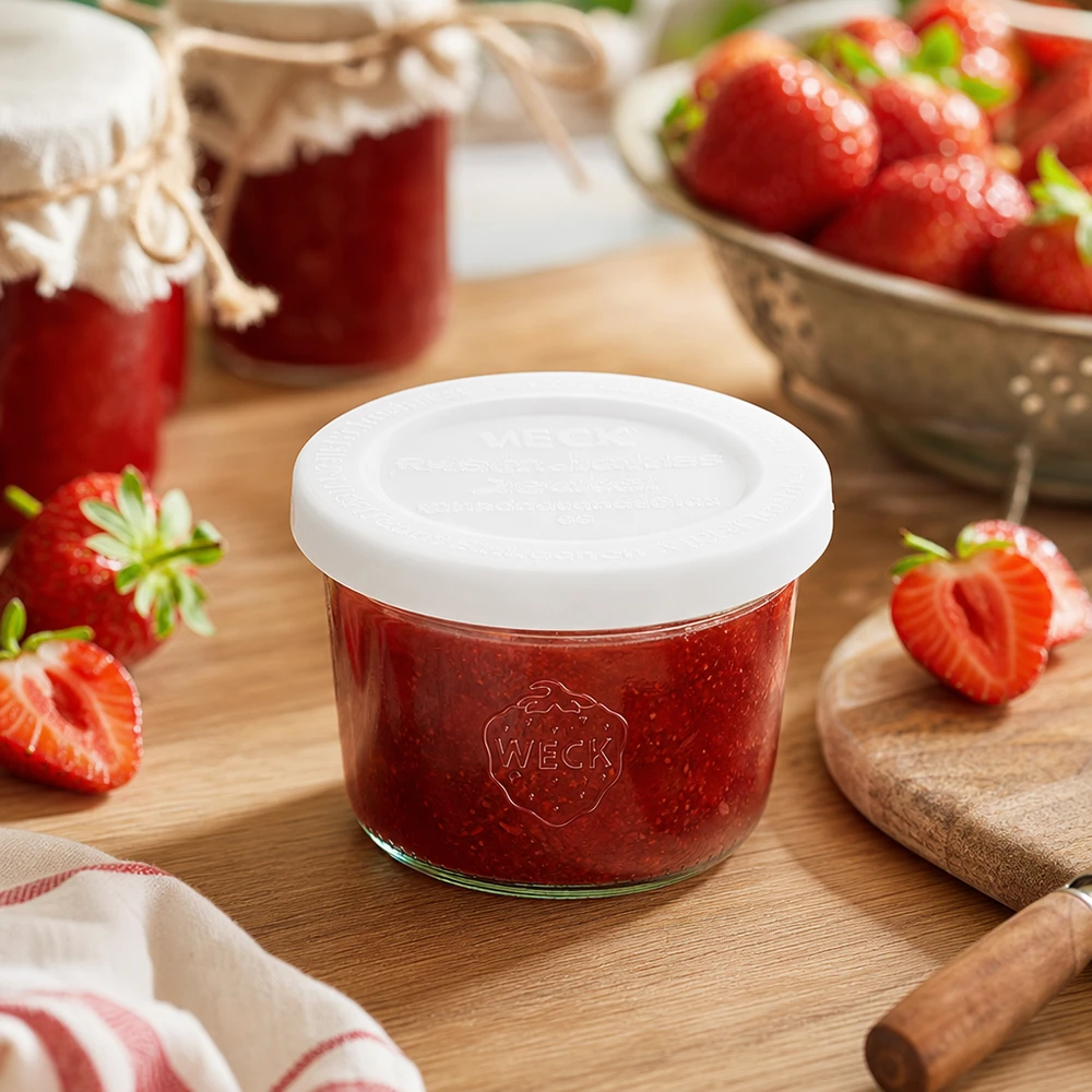 WECK RR60-pot van 80 ml met vershouddeksel WECK 80ml bekers RR60 gevuld met aardbeienjam op een houten plank omringd door verse aardbeien en jampotten